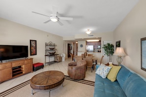 A spacious living room with a large flat-screen TV, a ceiling fan, and a dining area with wicker furniture.