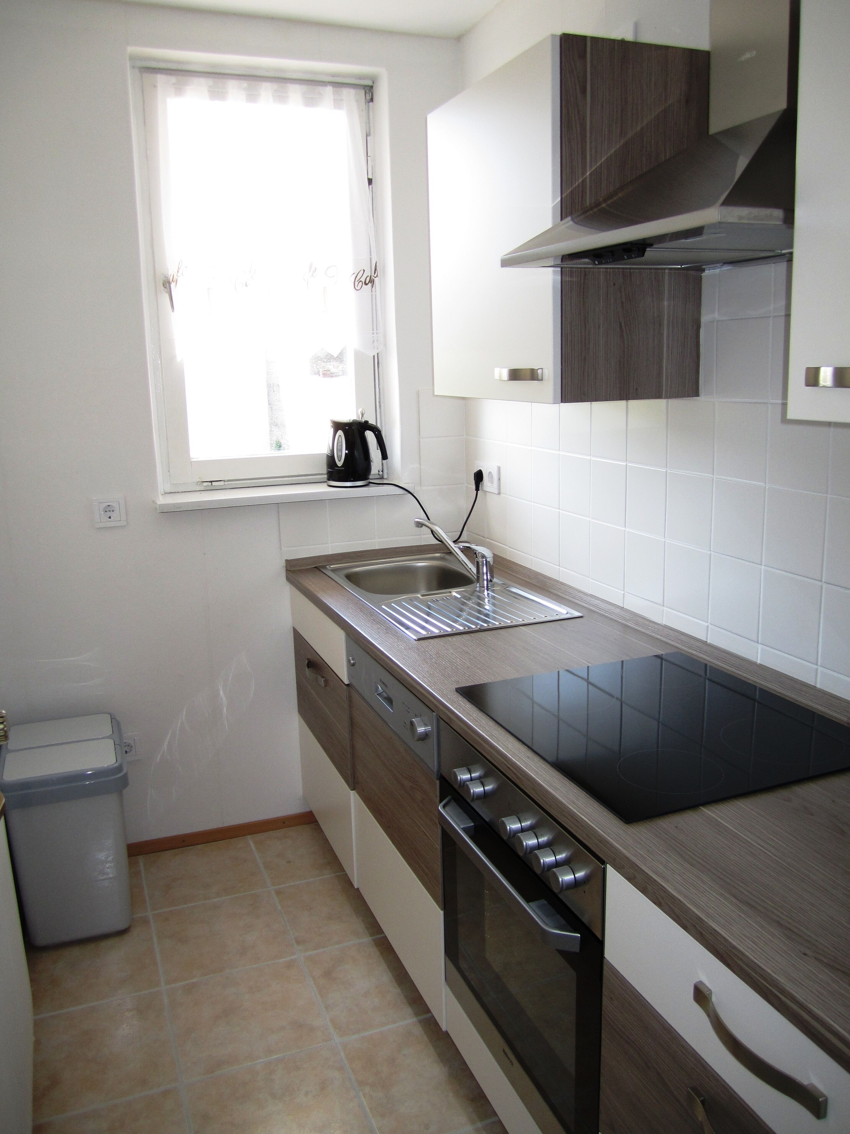 Cocina compacta con estufa integrada, un lavamanos y un ventanal con una tetera sobre la encimera.