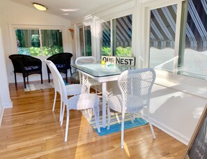 Un comedor luminoso con una mesa de vidrio y sillas de ratán blanco, un letrero que dice 'Our Nest', y ventanas amplias con vistas a la vegetación.