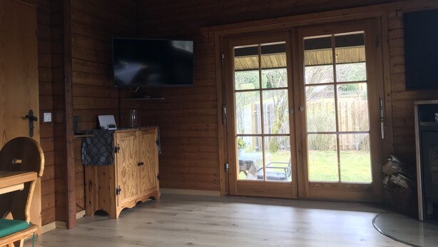 Interior de una cabaña de madera con una televisión montada en la pared, un armario de madera, un banco y un amplio ventanal que da a un espacio al aire libre.
