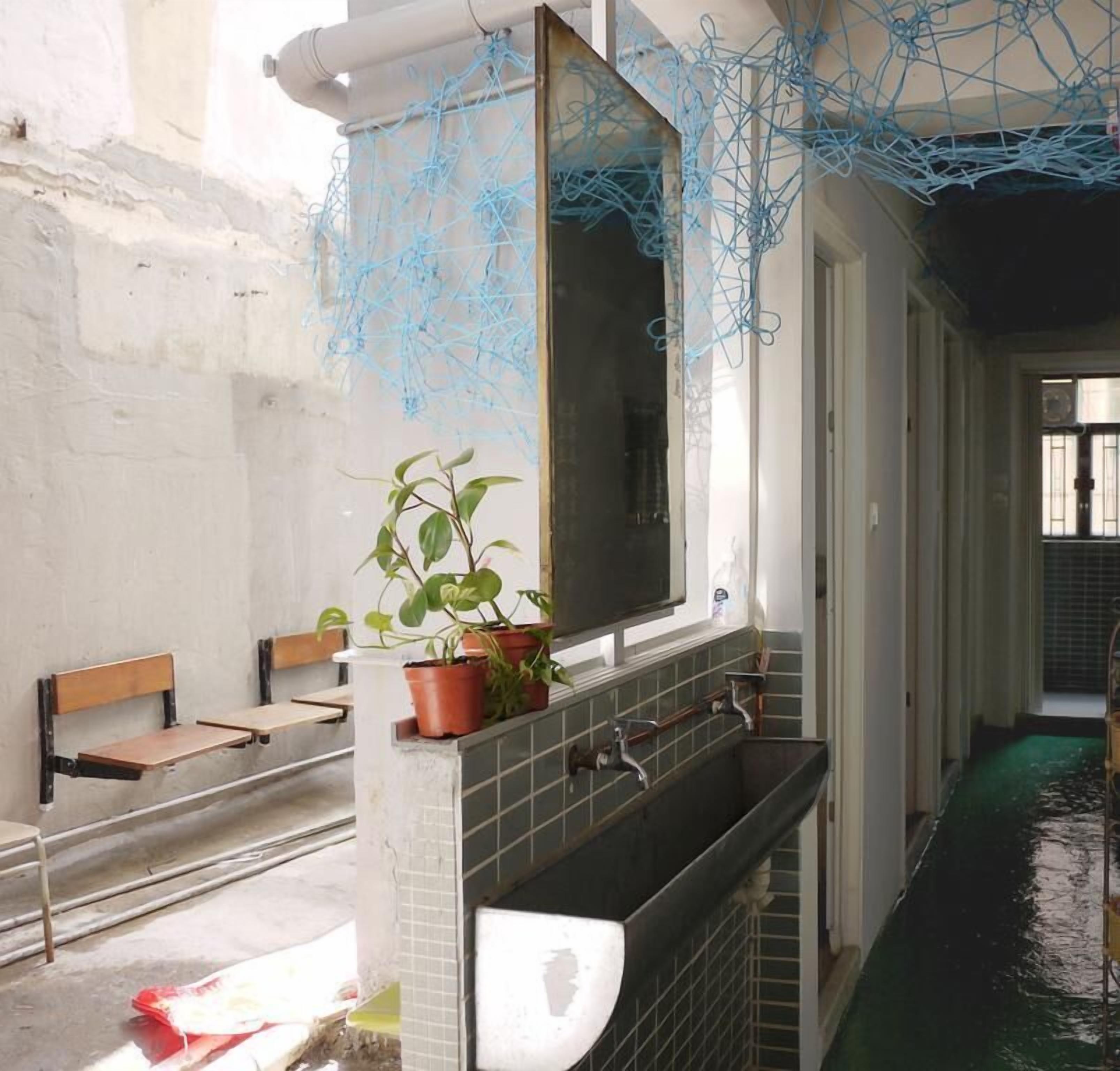 A room with a large mirror, a sink, and a bench. There are potted plants and a window with curtains.