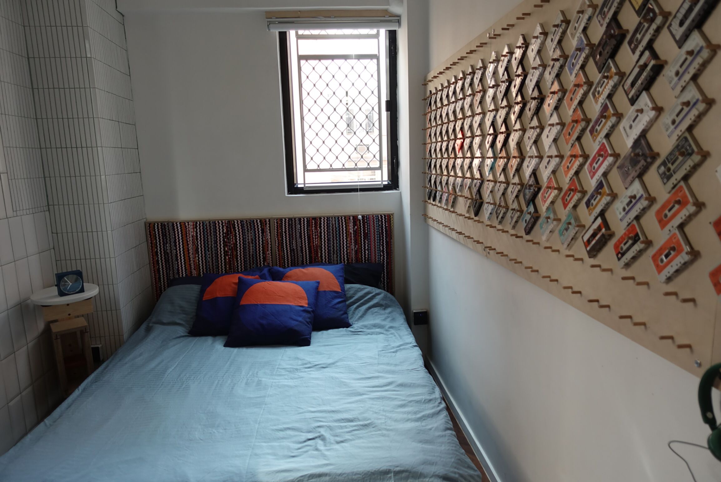 Ein Schlafzimmer mit einem Bett, einem Fenster, einem Wandregal mit Kassetten und einem kleinen Nachttisch mit einer Uhr.