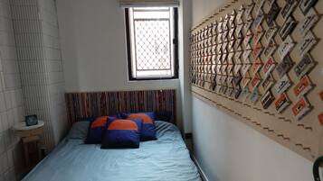 Ein Schlafzimmer mit einem Bett, einem Fenster, einem Wandregal mit Kassetten und einem kleinen Nachttisch mit einer Uhr.