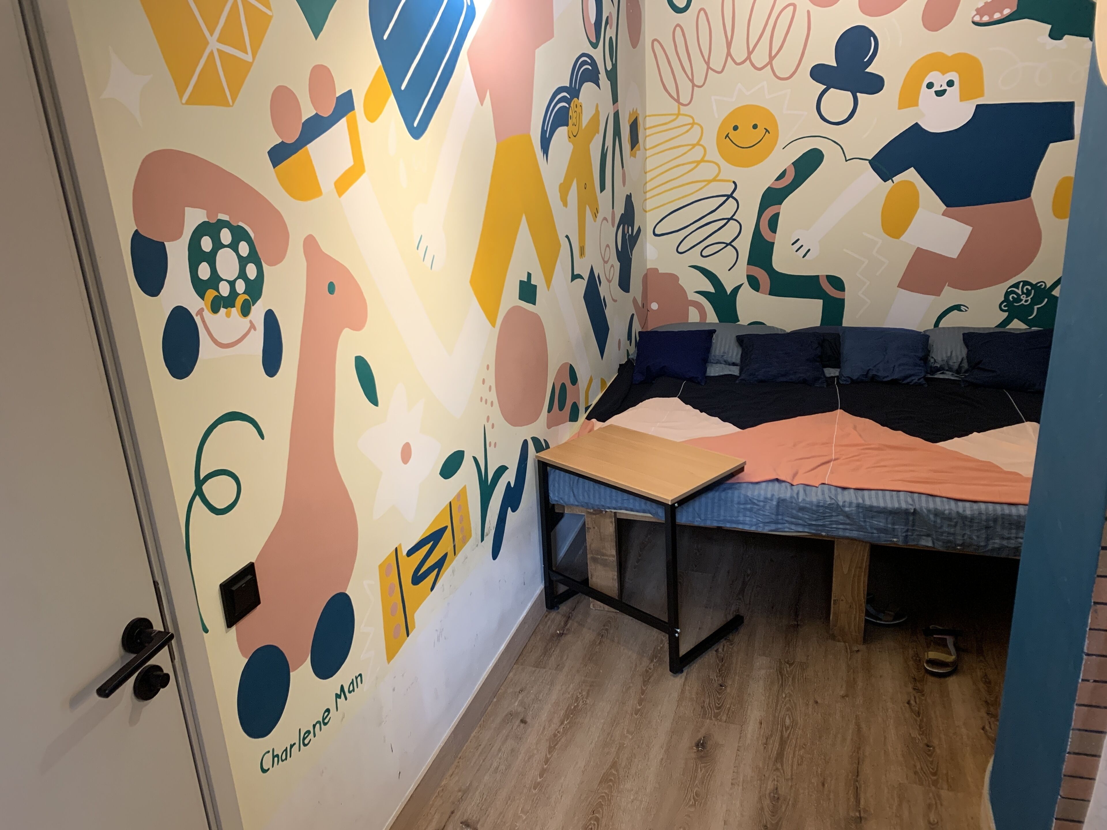A room with a colorful abstract mural, a bed with a blue cover, and a wooden floor.