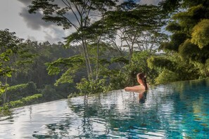 A person in a clear, infinity pool with a view of dense greenery and trees.