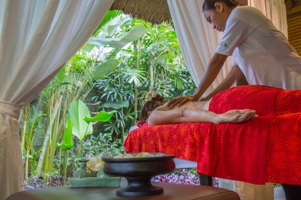A person receiving a massage in a room with a red massage table, surrounded by lush greenery.