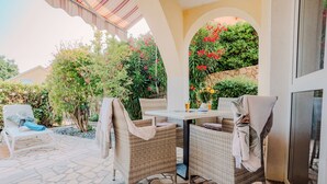 Ein sonnendurchfluteter Patio-Bereich mit Korbmöbeln, Steinboden und Blick auf einen Garten mit unterschiedlichen Pflanzen und Blumen.