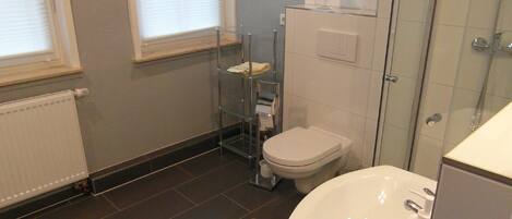 Une salle de bain avec des toilettes, un lavabo et une douche. Il y a une fenêtre et un radiateur.