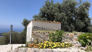Un petit bâtiment en pierre, doté d’un toit en tuiles, entouré de plantes en fleurs et d’un mur de pierre.