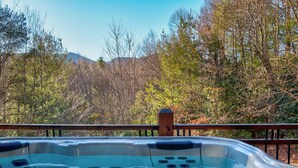 A hot tub with a wooden deck and a metal railing, surrounded by trees and a mountain in the background.
