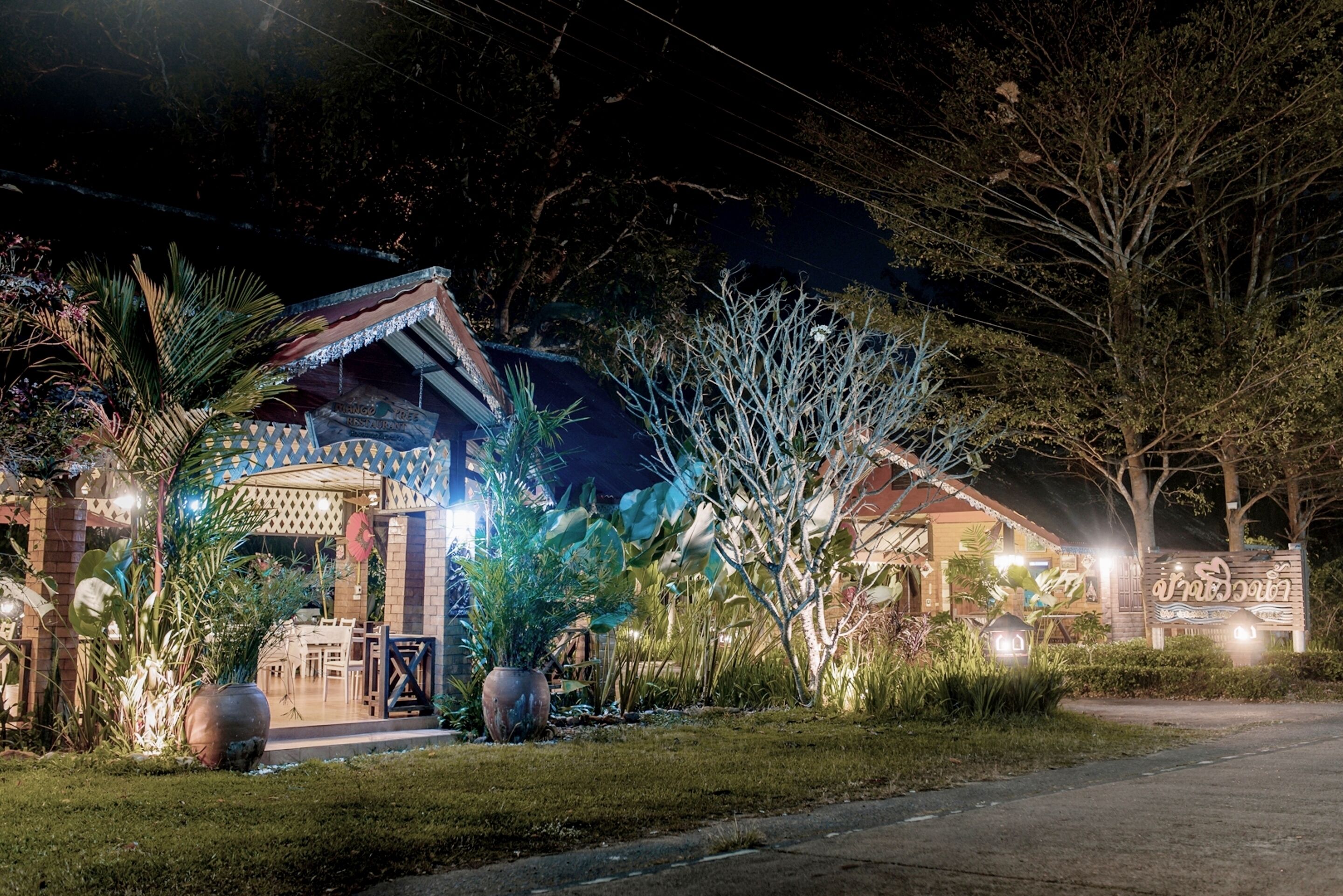 A well-lit outdoor area with a covered seating area, potted plants, and a pathway leading to a building with a sign.