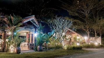A well-lit outdoor area with a covered seating area, potted plants, and a pathway leading to a building with a sign.