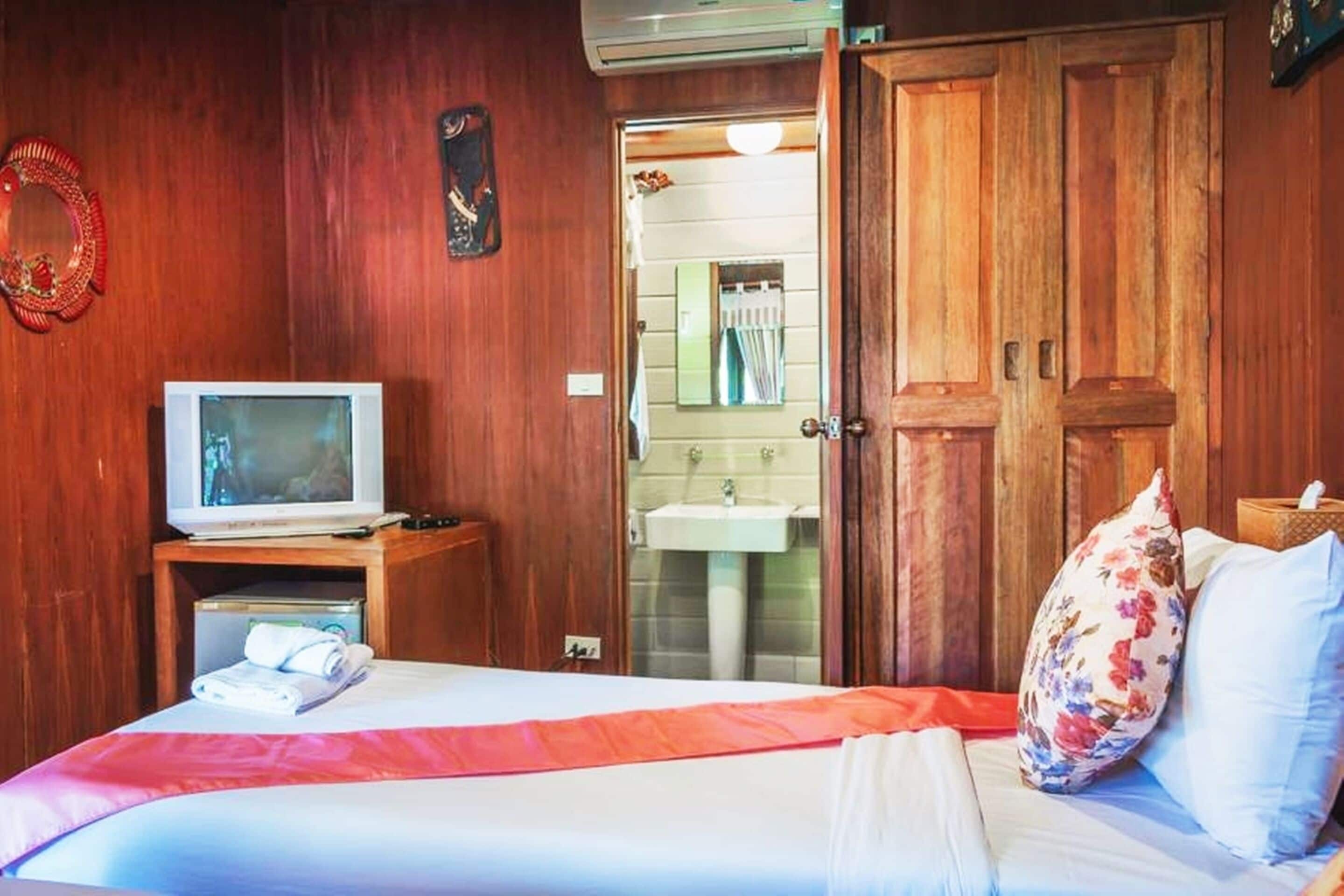 A hotel room with a wooden wall, a bed with white bedding and a red blanket, a television on a wooden stand, and a bathroom visible through an open door.