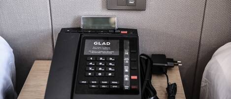 Habitación de hotel con un teléfono GLAD, un panel de control con opciones de iluminación y lectura, y un cable de alimentación en la mesita de noche.