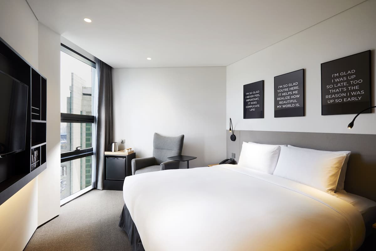 A modern hotel room with a large bed, a desk, a chair, and a TV. There are three motivational posters on the wall.