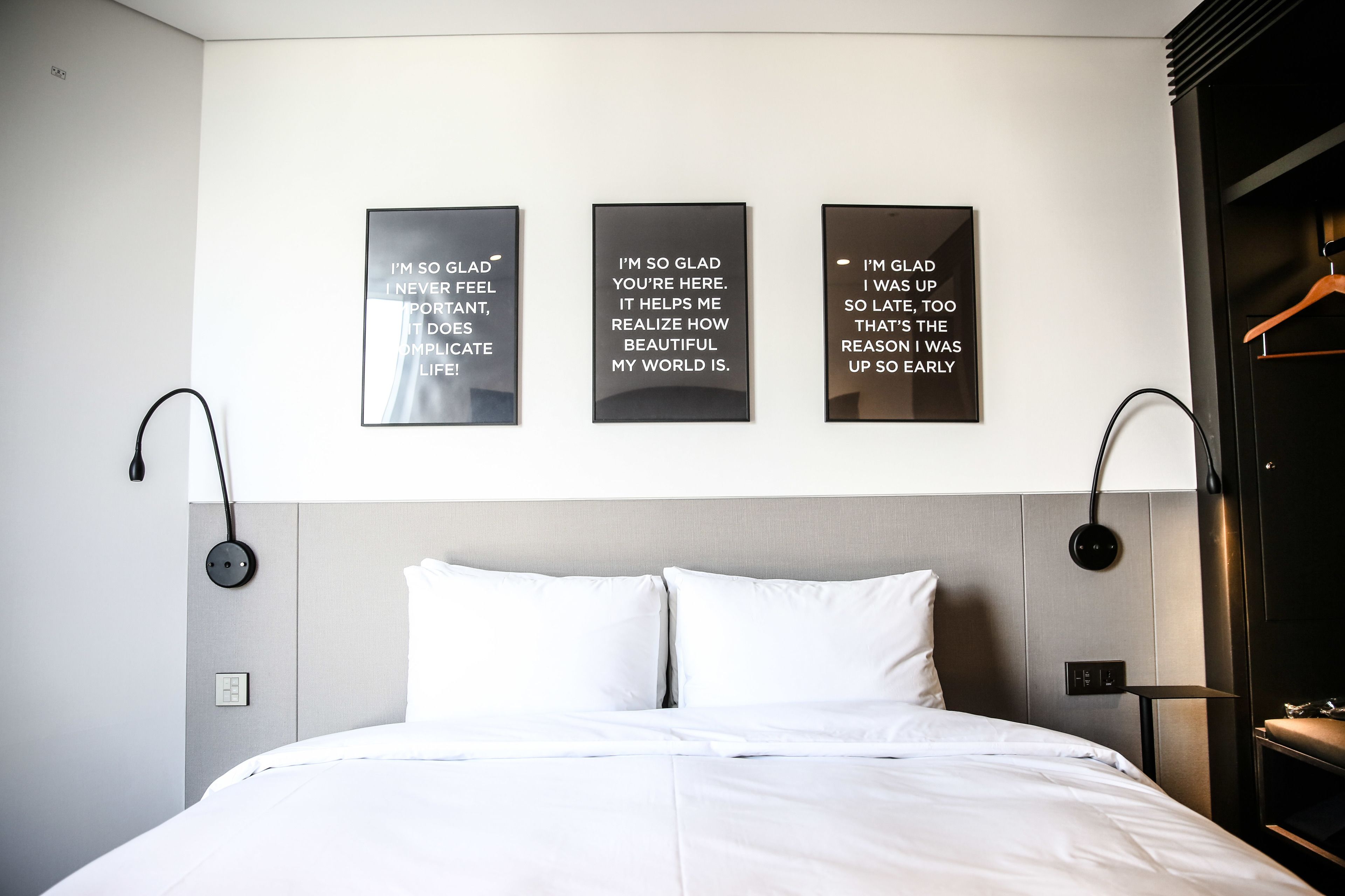A modern hotel room with a neatly made bed, a wall-mounted light fixture, and motivational quote posters.