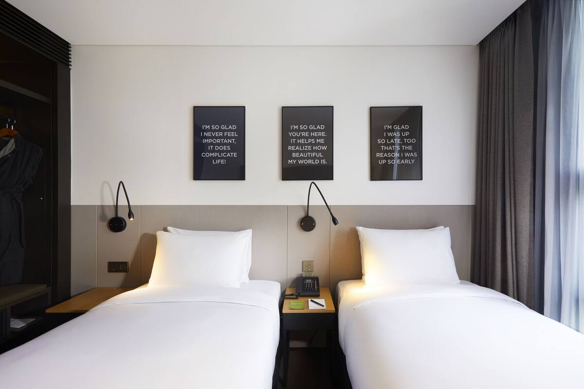 A hotel room with two beds, a bedside table with a phone and a lamp, and three motivational posters on the wall.