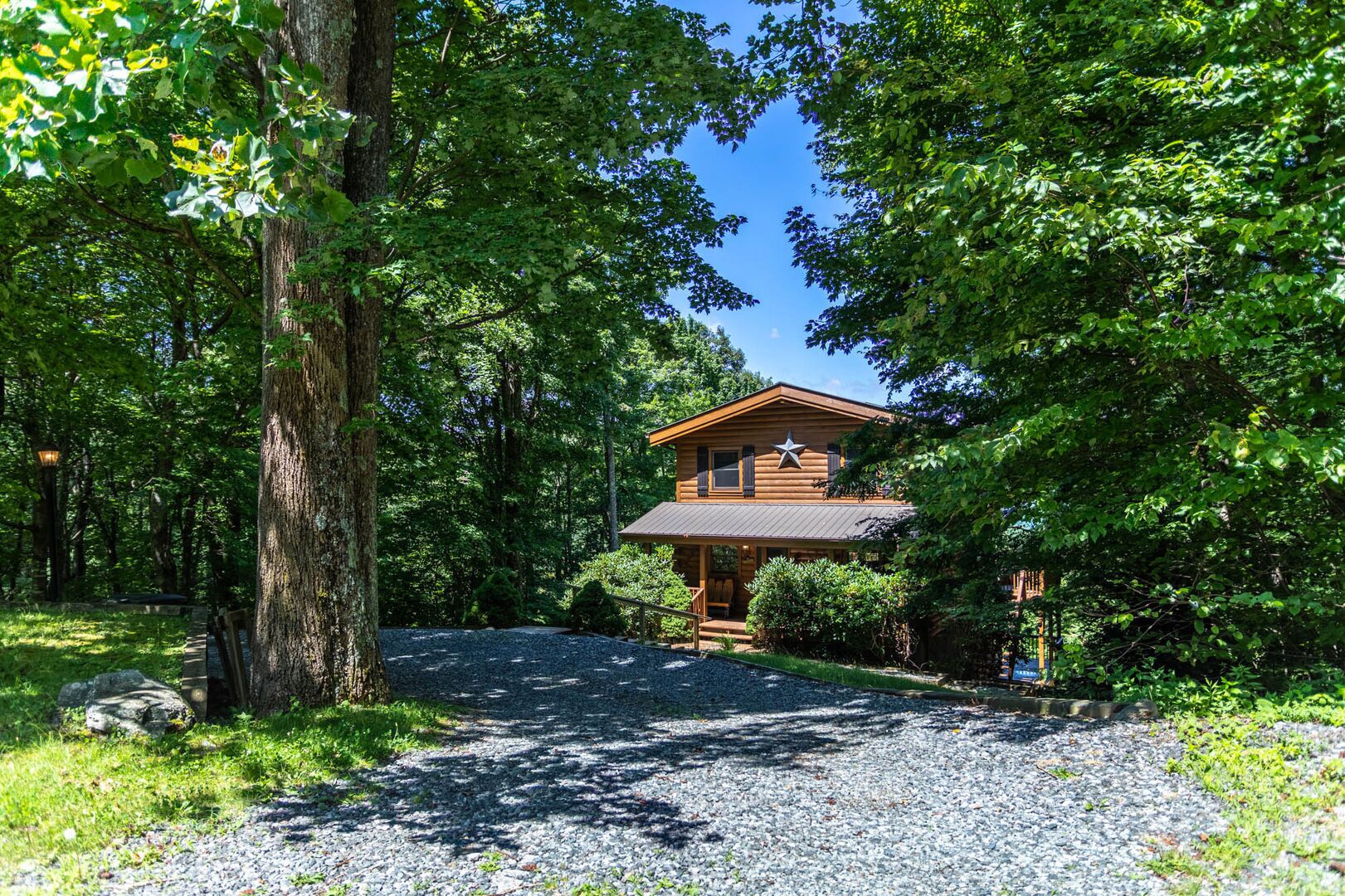 A two-story log cabin with a star on the front, surrounded by dense greenery and a gravel driveway.