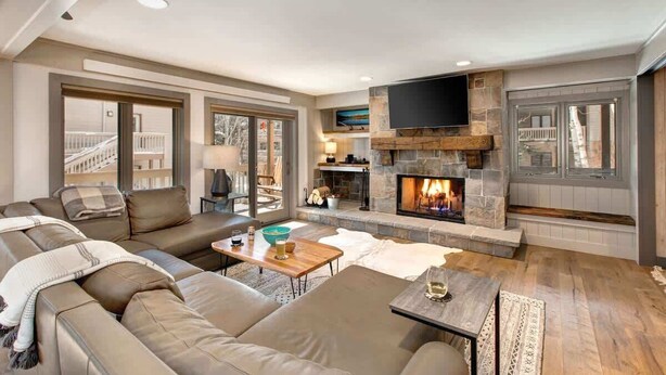 A modern living room with a stone fireplace, leather sofas, a flat-screen TV, and large windows.