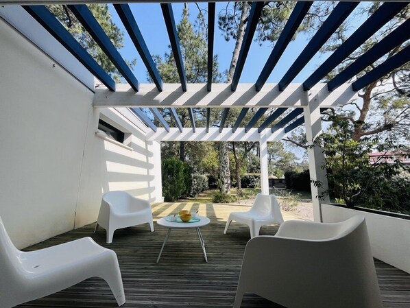 Un patio exterior con sillas blancas, una mesa pequeña con cuencos y una estructura de pérgola.
