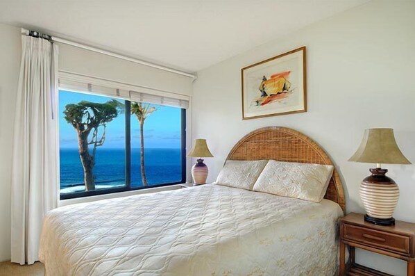 A bedroom with a large bed, two bedside lamps, a framed picture, and a view of the ocean through large windows. - Sealodge E-5, Offers dramatic views of the Pacific Ocean. (Princeville)