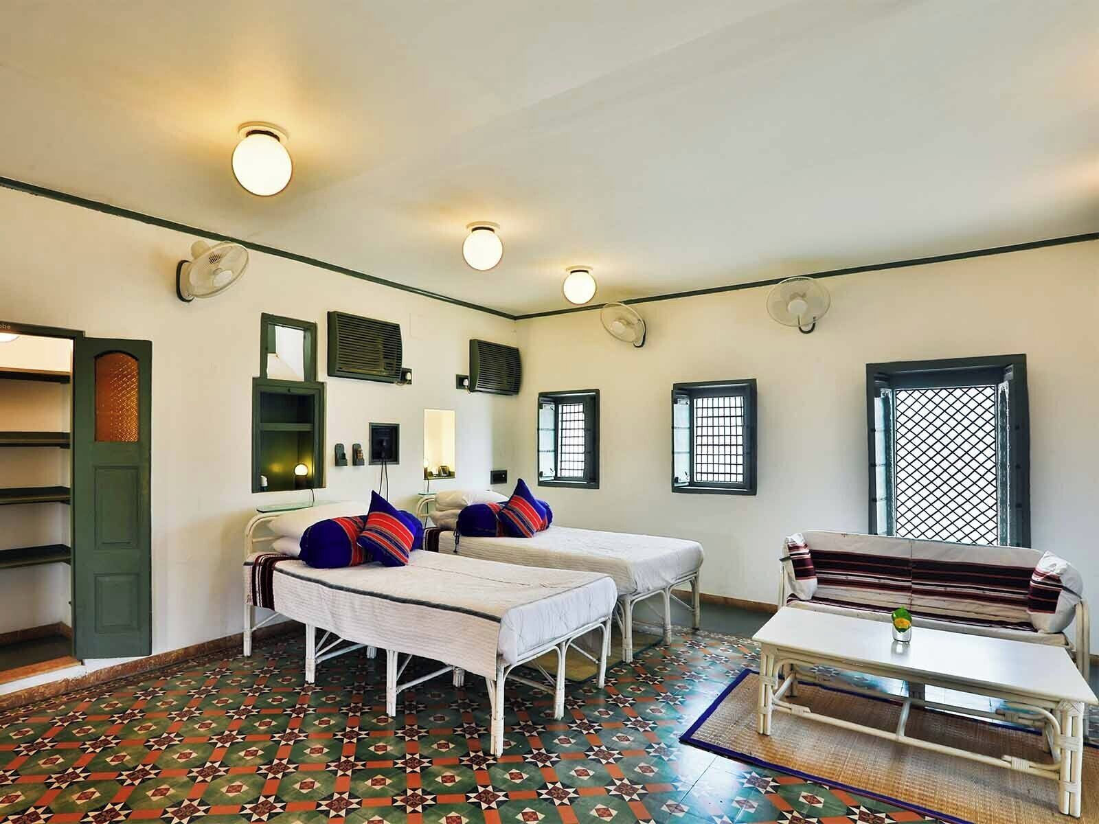 A room with two beds, a small table, and a bench. There are ceiling fans, a window with a metal grid, and a door with a green frame.