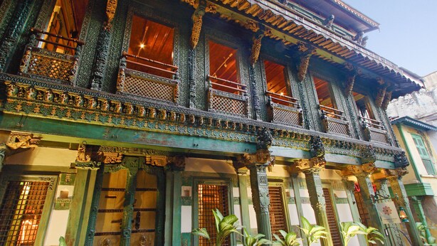 Un bâtiment traditionnel doté de balcons et de colonnes en bois richement décorés, avec des sculptures complexes et une porte en bois surmontée d’un portail en métal.
