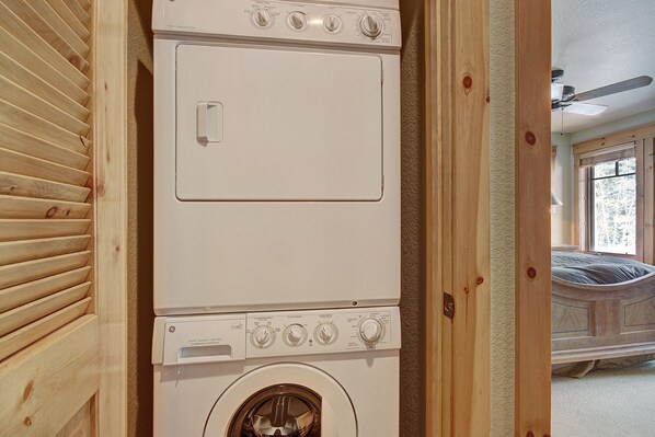 Lavadora blanca de carga frontal, compacta, en una habitación con suelo de madera, una cama y una ventana.