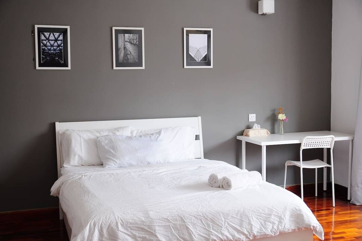 A neatly made bed with white linens, a small white desk with a chair, and three framed pictures on the wall.