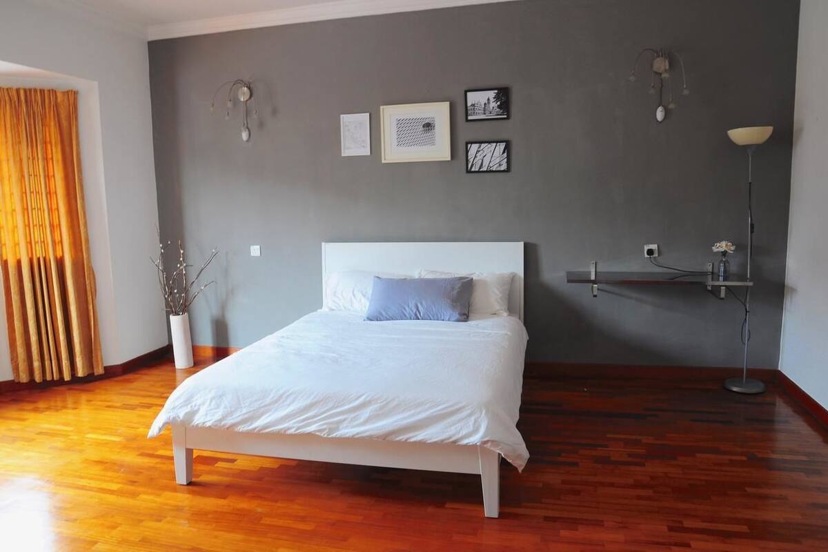 A bedroom with a white bed, a grey wall with framed pictures, a floor lamp, and a vase with branches.