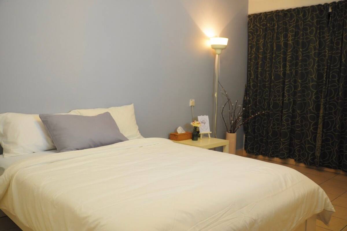 A neatly made bed with white and grey bedding, a bedside table with a lamp, and a patterned curtain.