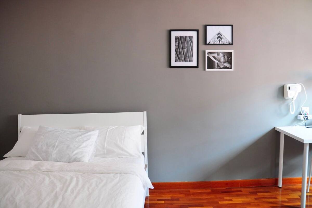A neatly made bed with white linens and pillows, a small white bedside table with a lamp, and a gray wall with three framed pictures.