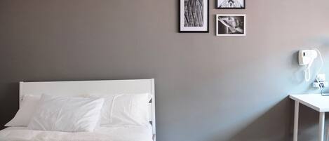 Una cama bien tendida con sábanas y almohadas blancas, una pequeña mesita de noche blanca con una lámpara, y una pared gris con tres cuadros enmarcados.
