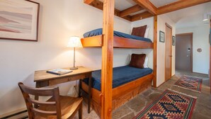 A dorm-style room with a bunk bed, a desk with a chair, a lamp, and patterned rugs on the floor.
