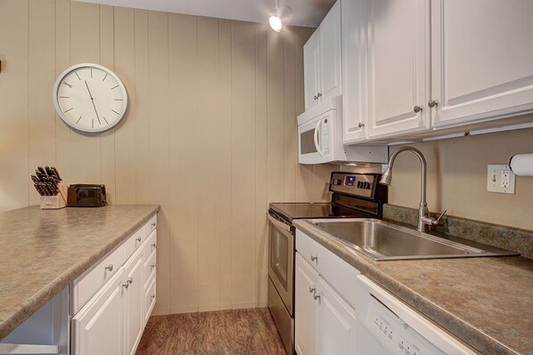 A compact kitchen with white cabinets, a stainless steel sink, a microwave, and a clock on the wall.