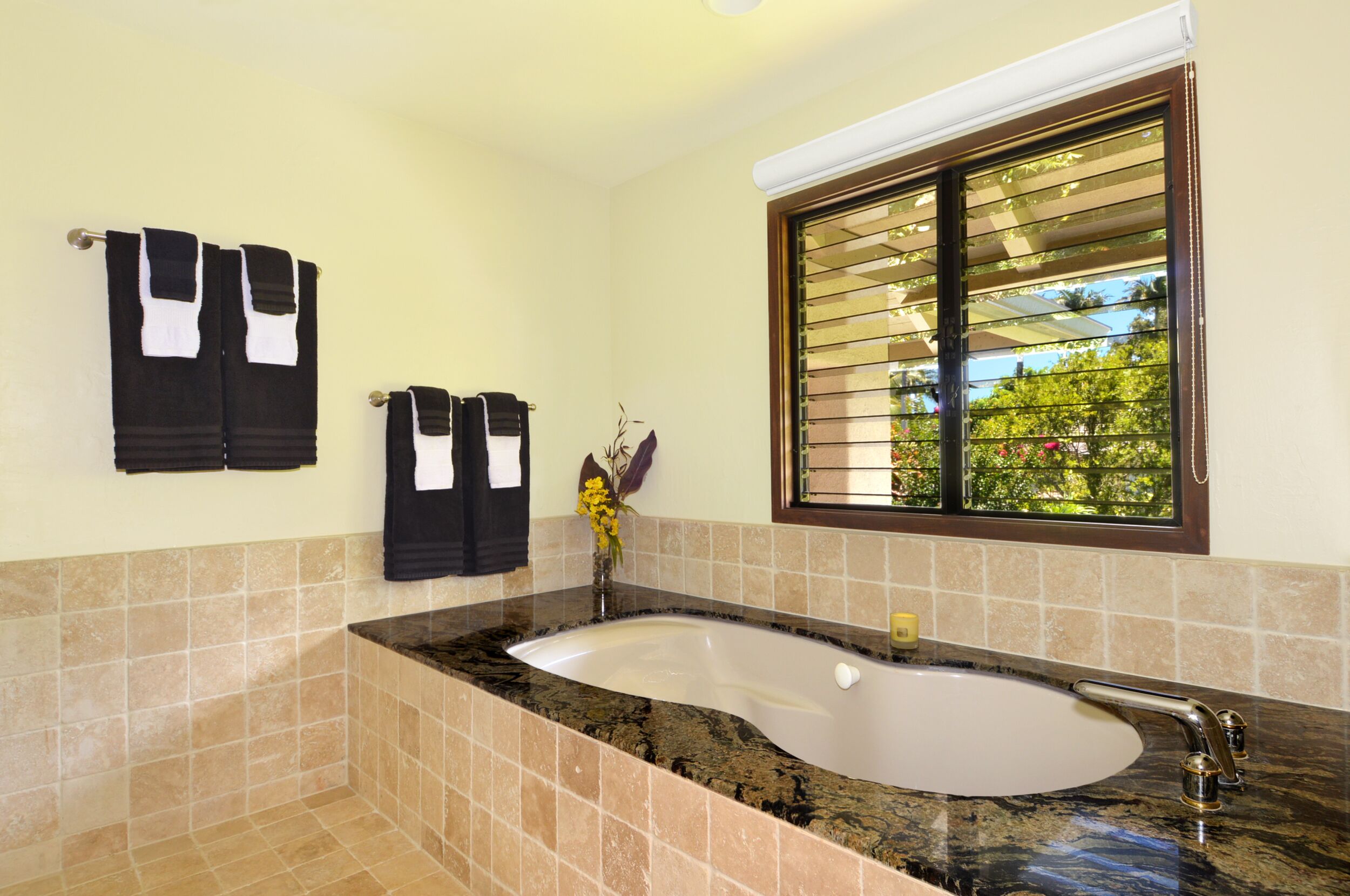 A bathroom with a large bathtub, beige tiled walls, and a window with blinds.