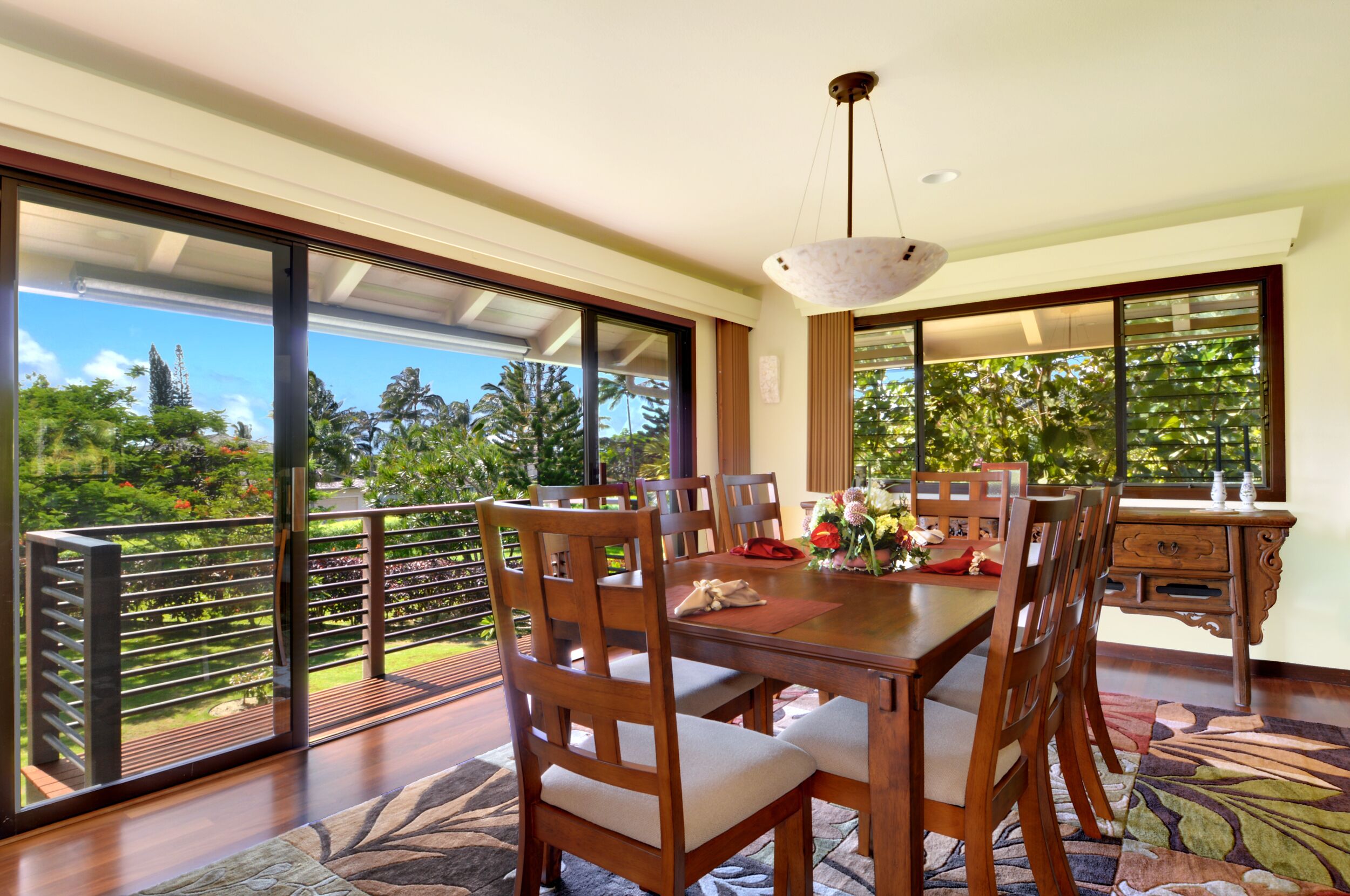 A dining area with a wooden table and chairs, a chandelier, and large windows offering a view of greenery.
