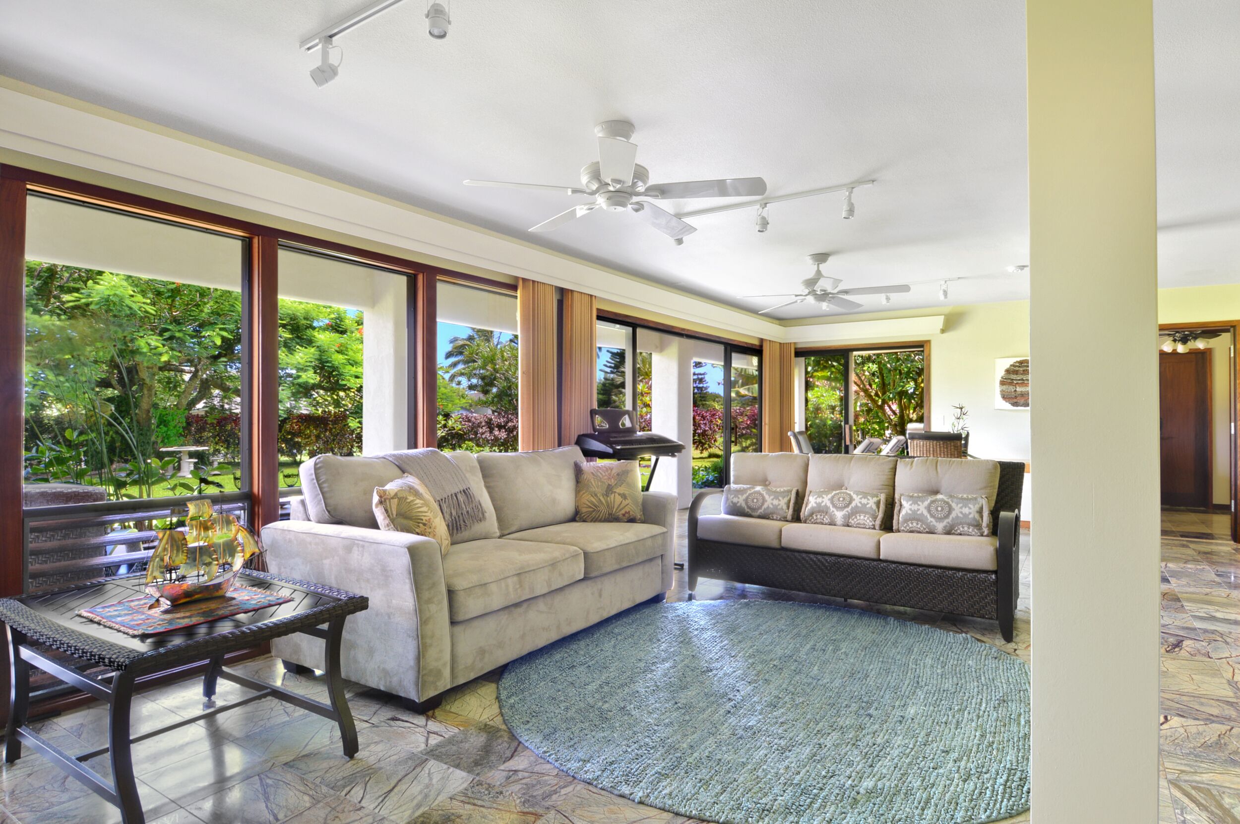 A spacious living area with two sofas, a coffee table, and a ceiling fan. Large windows offer views of greenery outside.