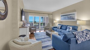 A living room with a blue sofa, a beige armchair, a coffee table, and a large window with a view of the ocean. - Sandy Key 121:  3 BR/2 BA Beachfront Condo, Perdido Key, FL. On-Site Rental Office, Sleeps 6 (Pensacola)