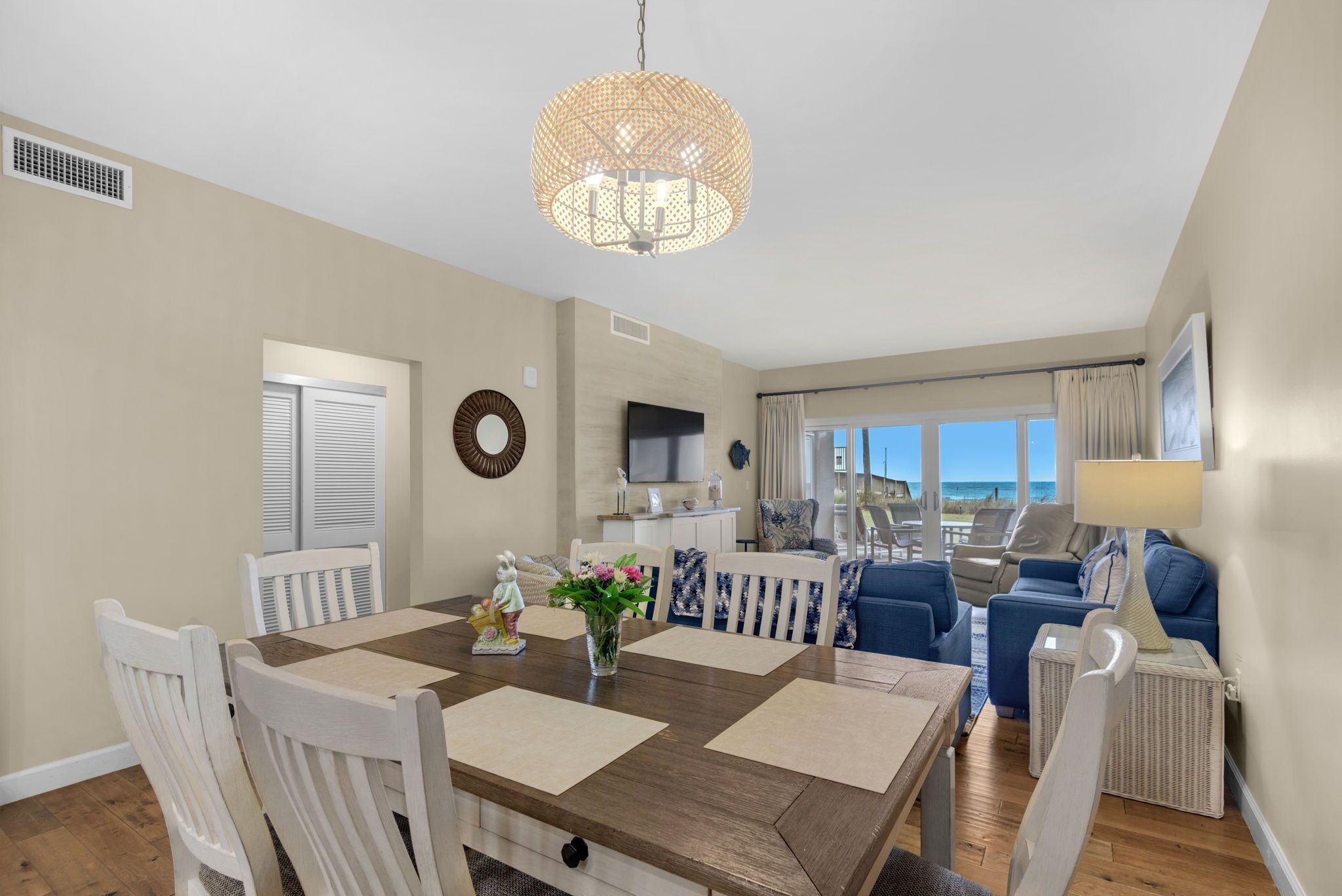 A spacious dining area with a wooden table and white chairs, a decorative hanging light fixture, and a view of the beach through large windows.