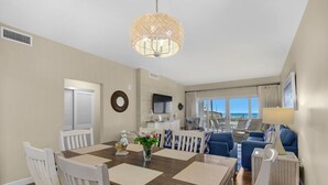 A spacious dining area with a wooden table and white chairs, a decorative hanging light fixture, and a view of the beach through large windows. - Sandy Key 121:  3 BR/2 BA Beachfront Condo, Perdido Key, FL. On-Site Rental Office, Sleeps 6 (Pensacola)