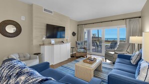 A living room with a large TV, a sofa, a chair, a coffee table, and a view through the glass doors. - Sandy Key 121:  3 BR/2 BA Beachfront Condo, Perdido Key, FL. On-Site Rental Office, Sleeps 6 (Pensacola)