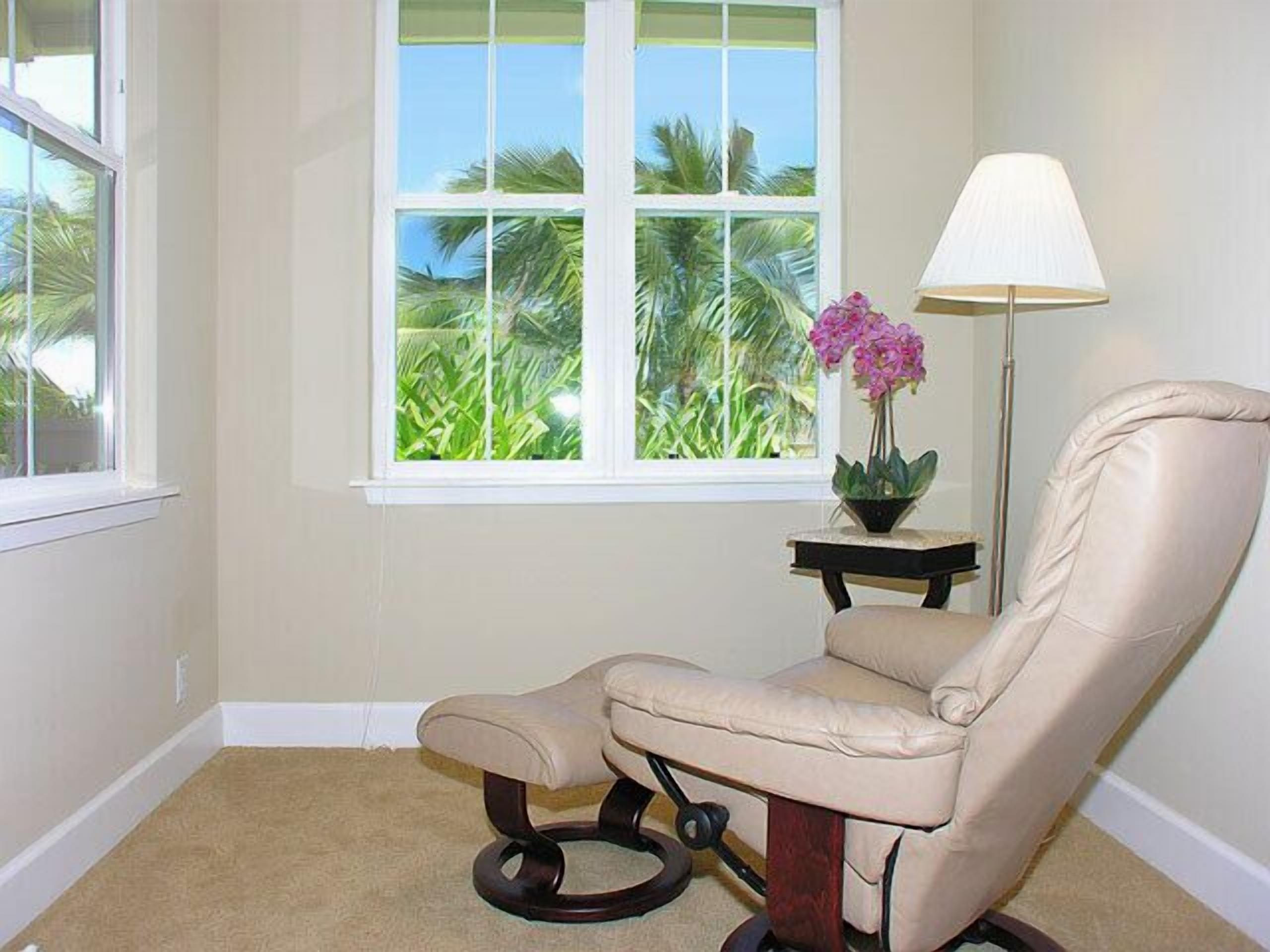A room with a beige leather chair, a small table with a lamp, and a window with a view of palm trees.