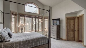 Un dormitorio con cama con dosel, cómoda, televisor y un amplio ventanal con vistas a árboles.