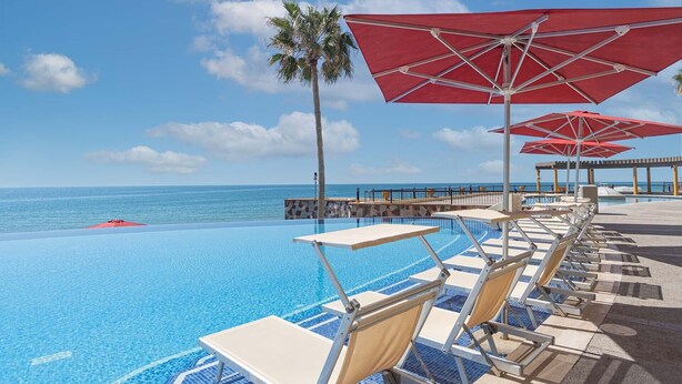 A poolside area with lounge chairs and red umbrellas, overlooking a clear blue ocean.