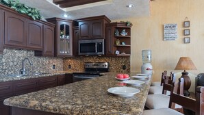 A kitchen with granite countertops, dark wooden cabinets, a microwave, stove, and a water dispenser. There are plates and apples on the counter.