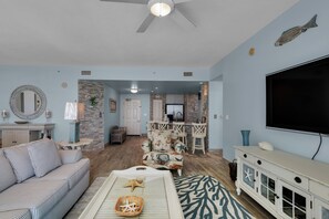 A living room with a sofa, coffee table, and a TV. There is a kitchen area in the background with a dining table and chairs. - 1402 -  Serenity by the Sea + Beach Chairs (Panama City Beach)