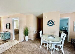 A dining area with a round table, white chairs, a potted plant, and a wall-mounted ship's wheel decoration.