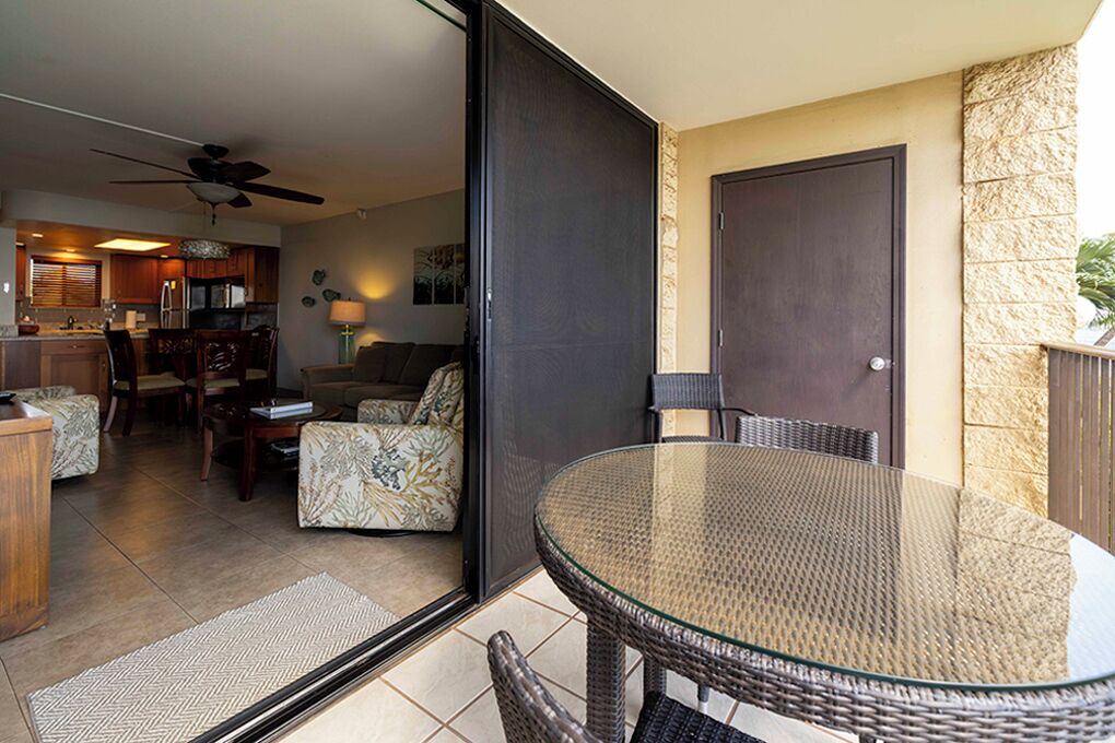 A glass-enclosed balcony with wicker furniture, a door, and a view of the kitchen and living area.