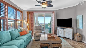 A living room with a teal sofa, patterned armchair, wooden coffee table, and a ceiling fan. - 101 West - Beachfront 3BR/3BA Corner Condo! (Gulf Shores)